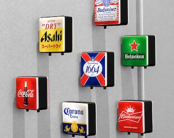 Könnte beinhalten: Eine Sammlung von sieben quadratischen, wandmontierten Leuchtschildern mit verschiedenen Getränkemarkenlogos, darunter Budweiser, Asahi Super Dry, Heineken, Kronenbourg 1664, Coca-Cola, Corona Extra und ein zweites Budweiser-Design mit Kronenlogo.