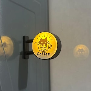 May include: A circular, wall-mounted LED sign with a yellow glowing face. The graphic features a black cat inside a coffee cup with the text WAKE CUP and Coffee printed in black. The sign is attached to a black metal bracket.