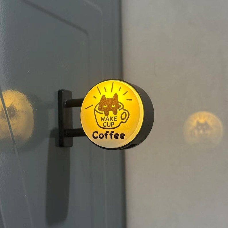 May include: A circular, wall-mounted LED sign with a yellow glowing face. The graphic features a black cat inside a coffee cup with the text WAKE CUP and Coffee printed in black. The sign is attached to a black metal bracket.