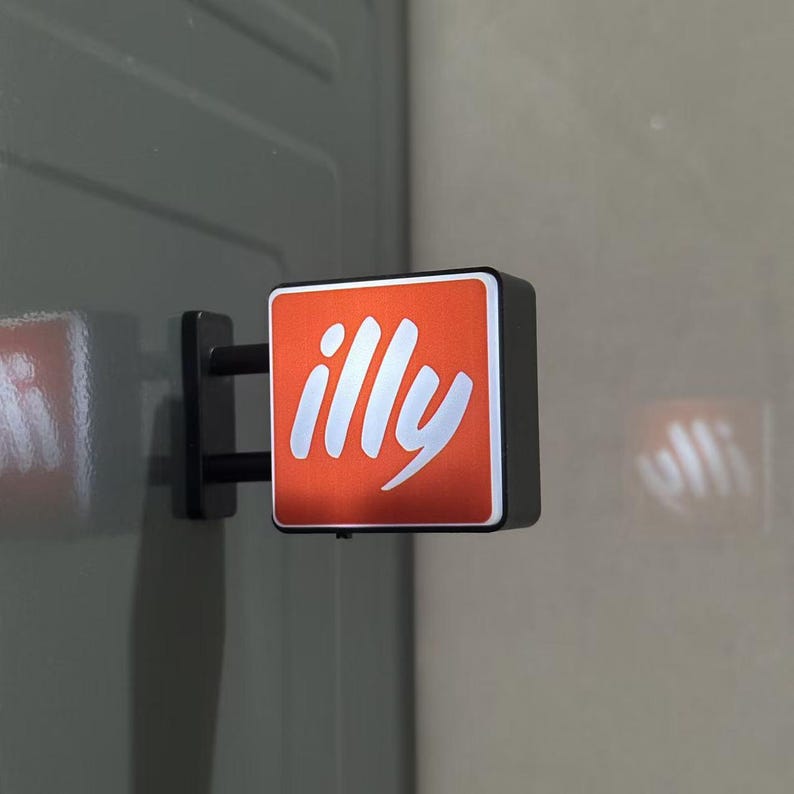 May include: A square, wall-mounted sign featuring a vibrant orange face with the white text illy in a stylized script font. The sign is attached to a black bracket and has a black frame, projecting outward from a flat, gray vertical surface.