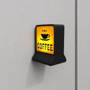 May include: A small, rectangular, black wall-mounted light fixture with a glowing yellow front panel. The panel features a black graphic of a coffee cup on a saucer with three small diamonds above it and the word COFFEE printed in black letters.