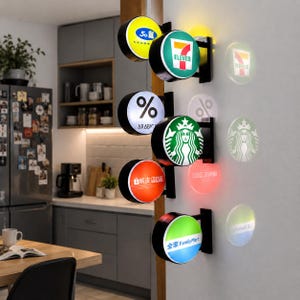 May include: A vertical wall-mounted display featuring five circular, illuminated signs with various brand logos including 7-Eleven, % Arabica, Starbucks, Shopee, and FamilyMart. Each sign projects a circular light pattern onto the adjacent white wall.