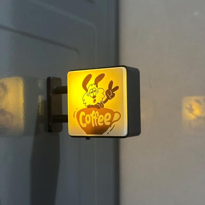 May include: A square, wall-mounted LED sign with a black frame and a glowing yellow face. The front panel features a dark brown graphic of a cartoon dog emerging from a coffee cup, making a peace sign, with the word Coffee written below.
