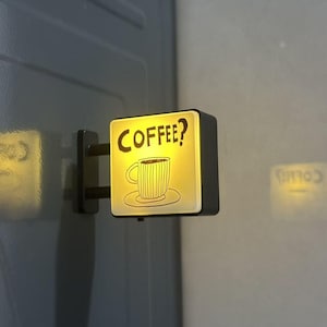 May include: A square, wall-mounted LED sign with a black frame and a glowing yellow face. The sign features a black line-art illustration of a coffee cup on a saucer with the text COFFEE? printed in a bold, hand-drawn style font above it.