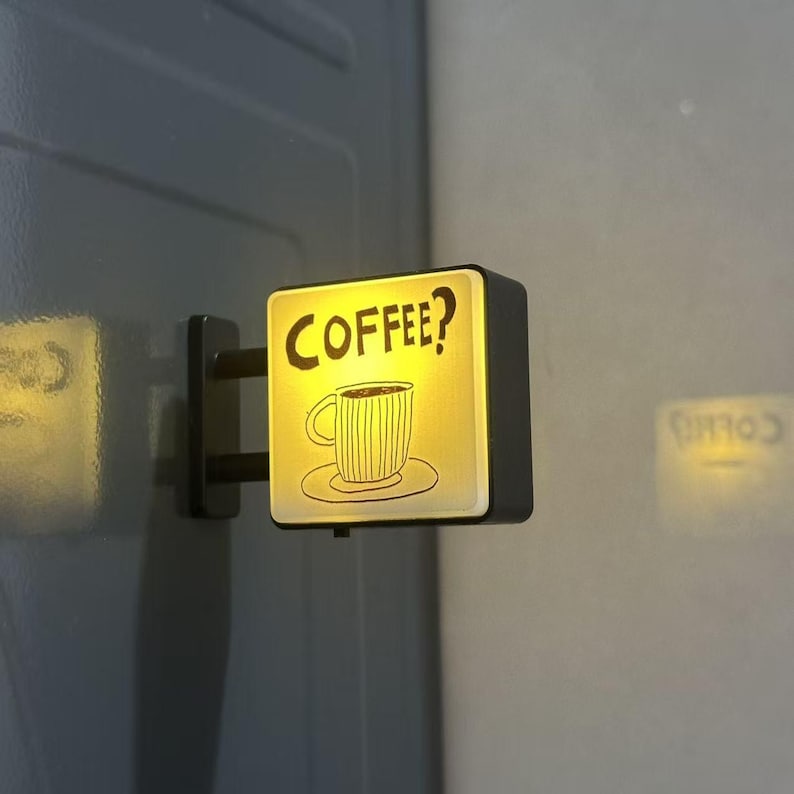 May include: A square, wall-mounted LED sign with a black frame and a glowing yellow face. The sign features a black line-art illustration of a coffee cup on a saucer with the text COFFEE? printed in a bold, hand-drawn style font above it.