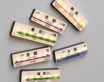 Könnte beinhalten: Sechs rechteckige, beleuchtete Schilder mit japanischen Bahnhofsnamen wie Shizuoka, Kyoto, Tokio, Osaka und Fukushima. Jedes Schild zeigt den Namen in japanischen Kanji und Englisch sowie farbige Richtungspfeile und das JR-Logo.