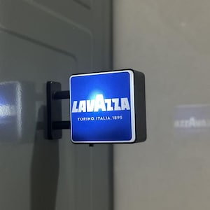 May include: A square, wall-mounted, illuminated sign featuring a blue background with the white text LAVAZZA and the smaller white text TORINO, ITALIA, 1895. The sign is attached to a black bracket and emits a bright light from its front face.