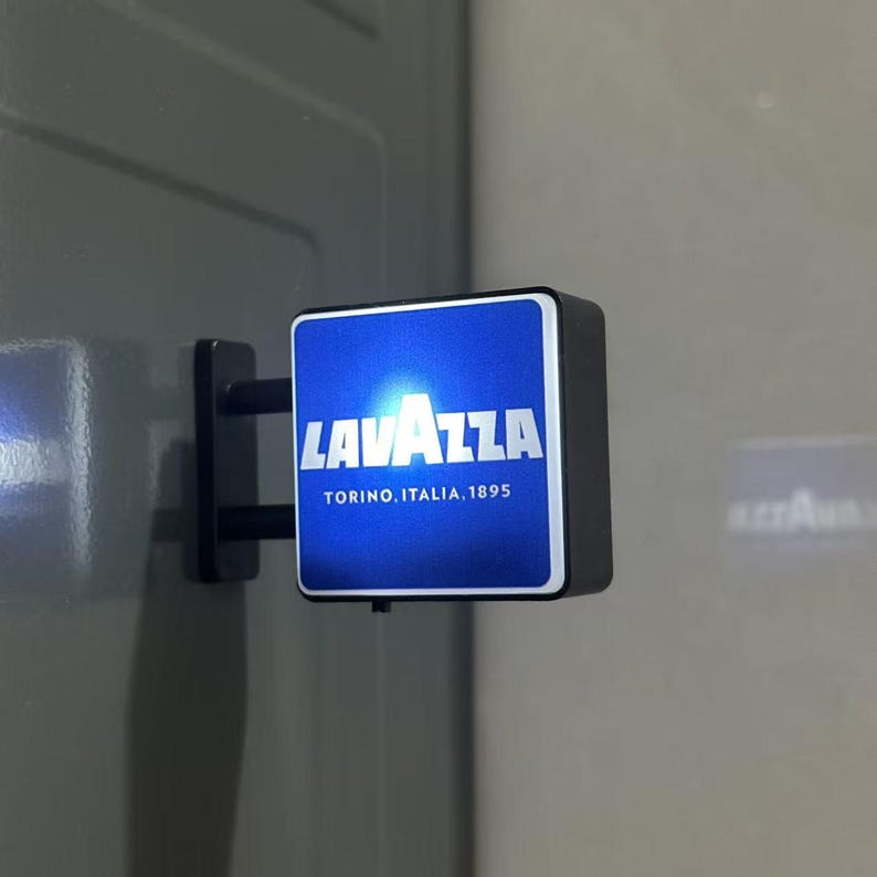 May include: A square, wall-mounted, illuminated sign featuring a blue background with the white text LAVAZZA and the smaller white text TORINO, ITALIA, 1895. The sign is attached to a black bracket and emits a bright light from its front face.