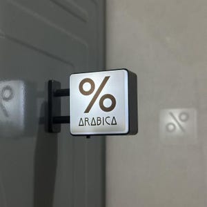 May include: A square, wall-mounted sign featuring a silver face with a brown percentage symbol and the text ARABICA printed in black. The sign is attached to a dark gray surface by a black metal bracket with two horizontal support bars.