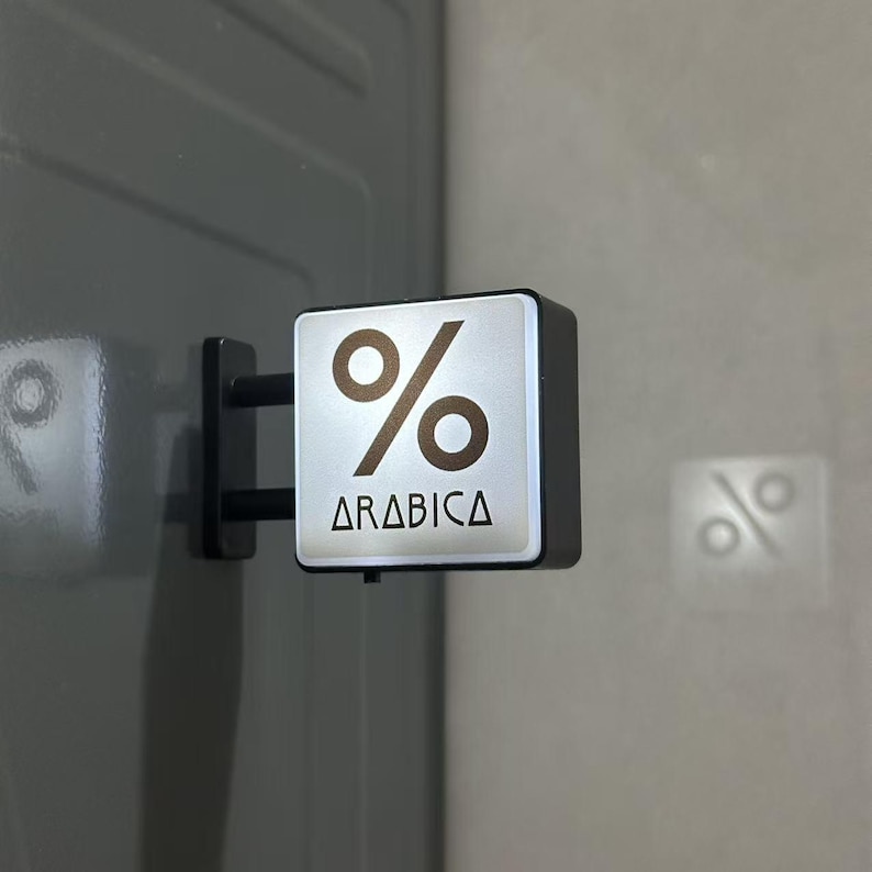 May include: A square, wall-mounted sign featuring a silver face with a brown percentage symbol and the text ARABICA printed in black. The sign is attached to a dark gray surface by a black metal bracket with two horizontal support bars.