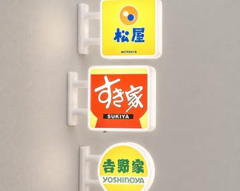 Könnte beinhalten: Drei beleuchtete Wandleuchtschilder. Das obere quadratische Schild ist gelb mit einem blauen Matsuya-Logo. Das mittlere quadratische Schild ist rot mit weißem Sukiya-Text. Das untere runde Schild ist gelb mit grünem Yoshinoya-Text und weißem Rand.
