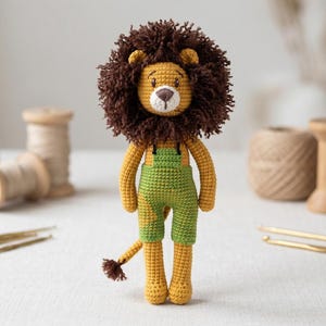 May include: A handcrafted crochet lion doll featuring a golden yellow body, a dark brown yarn mane, and green overalls. The lion has embroidered facial features, including eyes, a nose, and a mouth, with a small tail ending in a brown tuft.