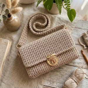 Könnte beinhalten: Eine beige gehäkelte Umhängetasche mit rechteckiger Klappe, langem passenden Schulterriemen und einem glänzenden goldfarbenen Metall-Druckknopfverschluss. Die Tasche ist in einem dichten, gleichmäßigen Maschenmuster aus beigem Garn gefertigt.