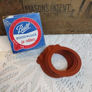 May include: A box of Ball Good Luck jar rubbers with a red, white, and blue label. The box is sitting on a white lace tablecloth with a stack of 10 brown rubber jar rings.