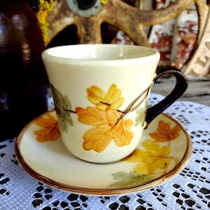 May include: Cream-colored teacup and saucer set with a leaf design in yellow, orange, and green. The cup has a black handle and sits on a matching saucer. The set is likely vintage or antique.