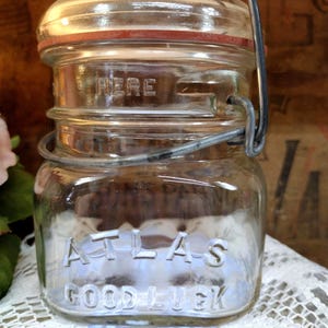 May include: Clear glass Atlas Good Luck jar with a metal wire closure and a glass lid. The lid has a red rubber seal. The word "HERE" is embossed on the jar. The jar is sitting on a white lace doily.