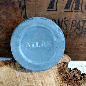 May include: A round, light blue metal lid with the embossed word "ATLAS" in the center. The lid is slightly weathered, suggesting age. The background includes a wooden surface and a portion of a wooden box.