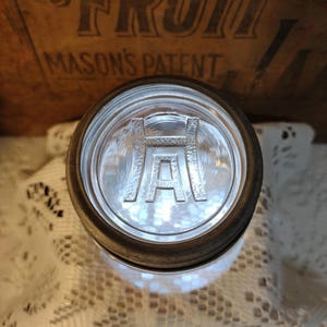 May include: A clear glass jar lid with a raised "H A" design in the center. The lid is surrounded by a metal ring. The jar lid is likely for preserving food. The background includes a wooden box with the text "MASON'S PATENT".
