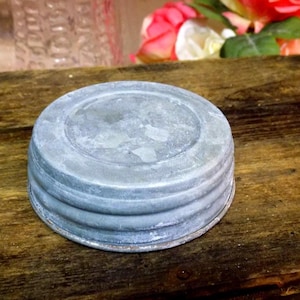 May include: A weathered, galvanized metal jar lid with a circular design. The lid has a series of concentric rings and a slightly domed top. The metal has a mottled, aged appearance, suggesting it is vintage or has been exposed to the elements. The background includes wooden planks and flowers.