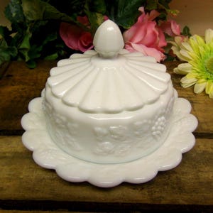 May include: A white, vintage butter dish with a scalloped edge base and lid. The lid has a fan-like design and a small, rounded handle. The dish features embossed grape and leaf patterns. A classic piece for serving butter.