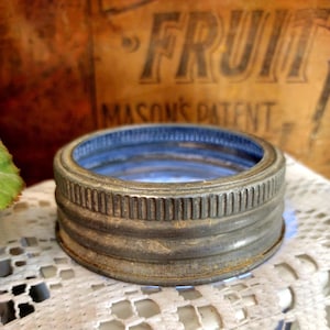 May include: A vintage, weathered metal Mason jar lid with a blue interior. The lid has a ribbed design and a slightly rusted appearance, suggesting age and use. The background includes a wooden crate with the word "FRUIT" visible.