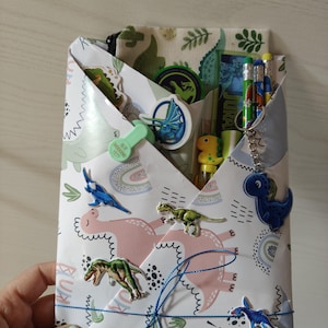 Könnte beinhalten: Ein weißer Papierumschlag mit rosa und grünen Dinosaurier-Illustrationen, der mit einer blauen Schnur zusammengebunden ist. Er enthält ein Federmäppchen mit Dinosaurier-Aufdruck, mehrere Stifte, einen Dinosaurier-Schlüsselanhänger und Aufkleber.