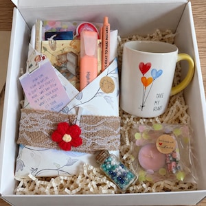 Puede incluir: Una caja de regalo blanca que contiene una taza de cerámica con asa amarilla y tres globos en forma de corazón, un resaltador color melocotón, una pequeña botella de vidrio con cuentas azules y blancas, una vela rosa y un paquete con una flor roja.