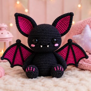 May include: A black and magenta crocheted bat plush toy with large pointed ears, wide black eyes, two small white fangs, and pink blush marks on its cheeks. The wings and inner ears feature a vibrant magenta color, while the feet have pink stitching.
