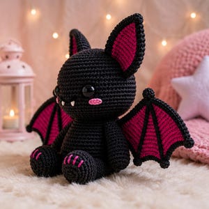 May include: A black crocheted bat plush toy with large black eyes, two white fangs, and pink blush marks on its cheeks. The bat features large pointed ears with pink inner sections and matching pink-accented wings and feet, all made of yarn.