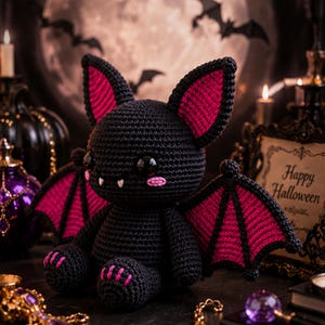 May include: A crocheted bat plush toy featuring a black body with vibrant pink accents on the inner ears, wings, and feet. The figure has large black glossy eyes, two small white fangs, and pink stitched cheeks, sitting in a seated position.