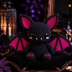 May include: A crocheted bat plush toy featuring a black body with vibrant magenta accents on the inner ears, wings, and feet. The figure has large black safety eyes, two small white fangs, and pink stitched cheeks, sitting in an upright position.