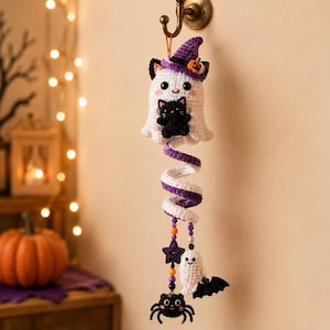Puede incluir: Una decoración colgante de ganchillo con un fantasma blanco que lleva un sombrero de bruja morado con una pequeña calabaza y sostiene un gato negro. Debajo, una espiral morada y blanca conduce a cuentas colgantes, una estrella morada, un fantasma pequeño, un murciélago y una araña negra.