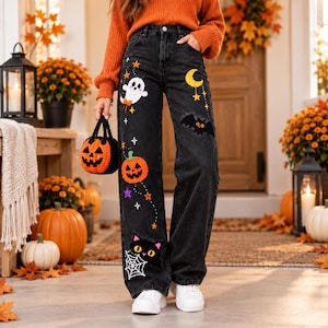 Peut inclure: Jean en denim noir orné de motifs brodés sur le thème d'Halloween, incluant un fantôme blanc, un croissant de lune jaune, une chauve-souris noire, une citrouille orange et un visage de chat noir avec une toile d'araignée blanche.