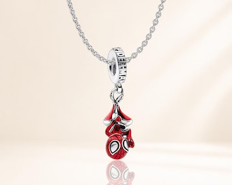 May include: A silver-toned necklace featuring a dangling charm of a red and black Spider-Man character. The charm is suspended from a circular silver bead engraved with the word SPIDER-MAN, which is threaded onto a delicate silver chain.