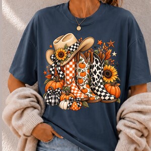 Peut inclure: Un t-shirt bleu foncé avec une impression graphique représentant un chapeau de cow-boy, trois bottes western, des tournesols et des citrouilles. Le chapeau et les bottes présentent des motifs à carreaux, peau de vache et citrouilles.