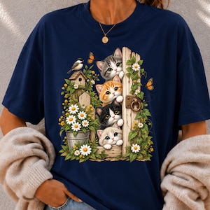 Può includere: Una t-shirt blu navy con una stampa grafica di quattro gattini che sbucano da dietro un telaio di porta in legno. La scena include una casetta per uccelli, un secchio di metallo pieno di margherite bianche, viti verdi e farfalle arancioni.