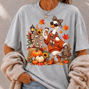 Peut inclure: Un t-shirt gris clair avec une illustration d'une oie blanche portant un bandeau léopard, des lunettes de soleil, une écharpe à carreaux, un pull orange et des bottes marron. L'oie tient un café et un sac à main léopard près d'un panneau en bois indiquant Fall VIBES Only, des citrouilles, des tournesols et des feuilles d'automne.