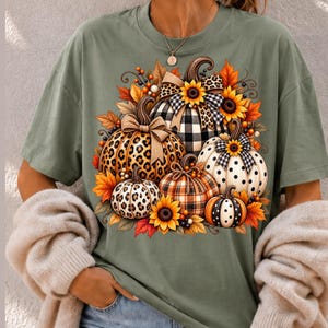 Peut inclure: Un t-shirt vert olive arborant une impression graphique de plusieurs citrouilles aux motifs variés, notamment léopard, carreaux vichy noir et blanc et pois, agrémenté de tournesols, de feuilles d'automne et de nœuds décoratifs.