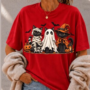Peut inclure: Un t-shirt rouge à manches courtes avec un motif de trois chats noirs déguisés pour Halloween. Un chat est momifié, un autre porte un drap de fantôme blanc, et le troisième porte un chapeau de sorcière et une cape orange. Citrouilles et chauves-souris.
