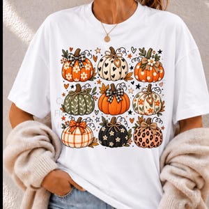 Peut inclure: Un t-shirt blanc à manches courtes avec une impression graphique de neuf citrouilles disposées en grille. Les citrouilles présentent divers motifs dont des carreaux, des pois, des taches de léopard, des cœurs et des fleurs, ornées de nœuds.
