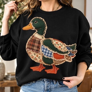 Peut inclure: Un sweat-shirt noir à manches longues orné d'une grande applique centrale représentant un canard colvert. Le canard est composé de divers empiècements en tissu, notamment des textures à carreaux, florales, vichy et velours côtelé.