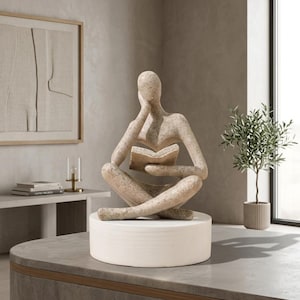 Pode incluir: Uma escultura de pedra bege salpicada de uma figura sem rosto sentada em posição de lótus enquanto segura um livro aberto. A figura apoia uma mão contra a cabeça e está montada sobre uma base cilíndrica branca e lisa.