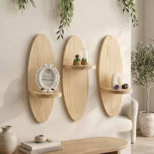 Puede incluir: Tres estantes de pared ovalados de madera clara con vetas naturales. Cada estante cuenta con una superficie horizontal plana unida al centro del panel trasero ovalado vertical. Los estantes sostienen pequeños objetos decorativos, incluido un marco blanco.