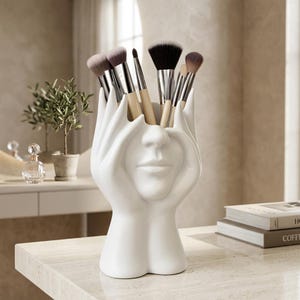 Peut inclure: Un porte-pinceaux en céramique blanche en forme de visage avec des mains entourant les côtés, rempli de divers pinceaux de maquillage aux manches en bois clair et aux poils foncés. La sculpture présente une finition mate et une couleur blanche.