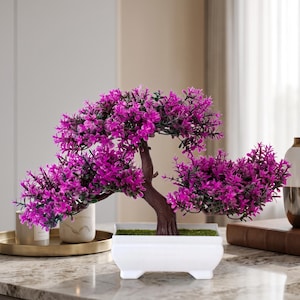 Könnte beinhalten: Ein künstlicher Bonsai-Baum mit leuchtend magentafarbenen Blättern und einem strukturierten braunen Stamm, gepflanzt in einem rechteckigen weißen Keramiktopf, der mit grünem Kunstmoos gefüllt ist.