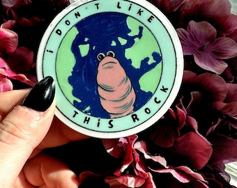 May include: A round sticker with a cartoon earth design and the text "I DON'T LIKE THIS ROCK." The earth is blue and green, with a pink worm-like creature in the center. The sticker has a light green border.