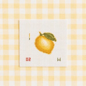 May include: A square white cross-stitch fabric piece featuring a yellow lemon with a green leaf in the center. The design includes the vertical bar symbol, the number 02 in red, and the letter W in green stitched on the white canvas surface.