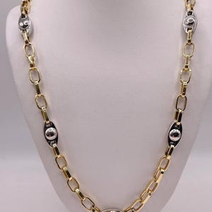 Puede incluir: Un collar de cadena bicolor con eslabones rectangulares alargados de color dorado intercalados con estaciones ovaladas de color plateado. Cada estación plateada contiene una esfera pulida central, creando un patrón repetitivo.