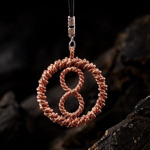 May include: A copper wire pendant shaped like an infinity symbol enclosed within a circular frame. The pendant is crafted from twisted copper wire and hangs from a black cord secured by a silver-toned metal bail at the top of the circular frame.