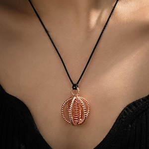 May include: A rose gold-toned pendant featuring a spherical, cage-like design made of twisted metal wire, suspended from a thin black cord. The pendant has a central coiled element surrounded by multiple curved, textured wire loops.